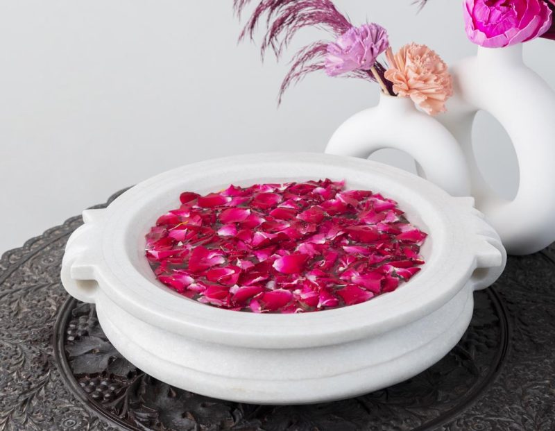 Traditional white Urli Candle Floater brimming with vivid pink rose petals, accompanied by modern white sculptures, merging classic and contemporary decor styles.